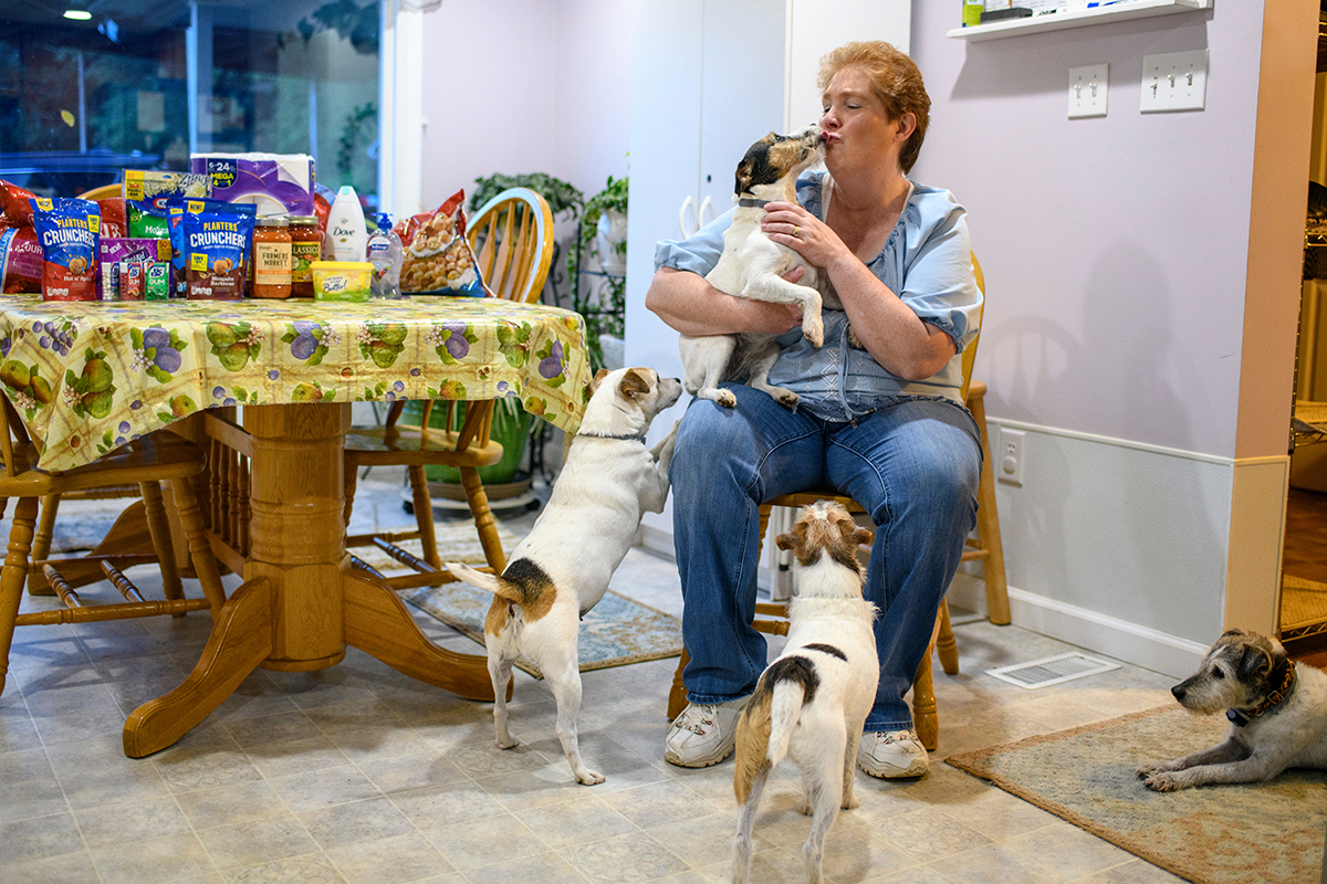 A woman sits in a kitchen and holds one of her three dogs.