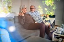 A senior citizen couple sit on their couch while discussing something.