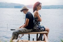 An unhappy couple sit on a dock overlooking the water.