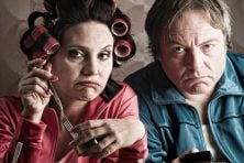 A couple eat out of tin cans while looking unhappy. The woman's hair is in curlers.