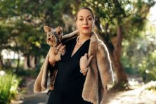 A woman wears a black dress, pears and a fur coat. She's also holding a small dog.