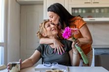 An adult daughter brings flowers to her mother. Her mother is sitting at the kitchen table.