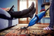 A man lifts his legs up so his wife can vacuum underneath him.