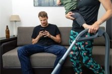A man plays on his phone as his wife cleans with their child in their arms.