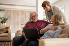 Grandparents sit on a couch with a laptop as their grandson hovers over them listening to what they have to say.