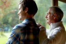 A grandson and grandfather stand next to a window.