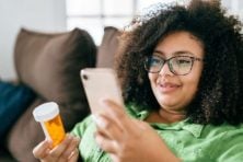 A woman holds a prescription in her and while looking at her phone with a smirk on her face.