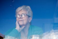 An elderly woman looks out the window of her home with a look of loneliness.