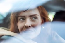 A woman stares into the distance while resting her head on her hand that's on her steering wheel inside her car.