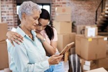 A daughter hugs her elderly mother as they tenderly look at a photo with moving boxes all around them.