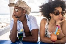 A couple look unhappy on a beach vacation with mixed drinks next to them.