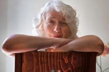 A woman sits backwards on a chair while lookingat the camera with a slight smirk.