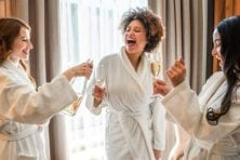 Three women wear hotel robes and celebrate with champagne in a hotel room.