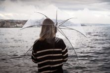 A woman holds a see through umbrella while standing near water on a cloudy and cold day.