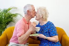 A man kisses his wife on the nose as they have tea together on their yellow couch.