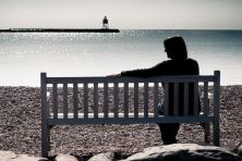 A woman looks at an empty seat on a park bench as she sits near the ocean with a lighthouse in the background.