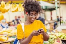 A woman smiles as he holds a basket of produce and looks at her phone.