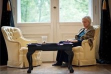 A woman looks sad sitting in a chair with an empty one next to it. This is meant to represent a widow.