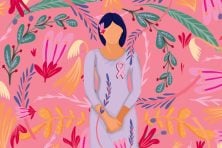 A woman stands against a pink background with flowers as she has the breast cancer awareness symbol on her chest.