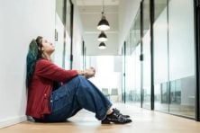 A woman sits down in a hallway in an office building. She looks up toward the ceiling with an upset expression on her face. She's wearing a red sweater and blue loose-fitting jeans.