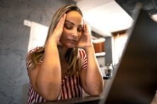 A woman looks stressed as she brings her hands to her temples while looking at her laptop.