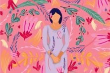 A woman stands against a pink background with flowers as she has the breast cancer awareness symbol on her chest.