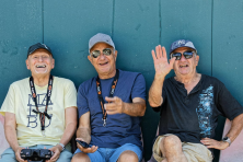 Three retired men laugh as they hold remote controllers outside on a bench.