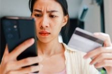 A woman has a pensive look on her face as she looks at her phone with her credit card in the other hand.