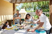 A group of people with children set the table for lunch together outside underneath a pergola.