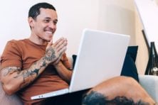 A man looks excited with his white laptop on his lap. The man has an armful of tattoos and is smiling with his hands clasped together.