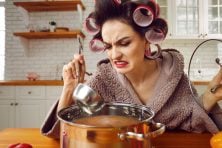 A woman looks grossed out as she stirs a pot of soup with curling rolls in her hair.