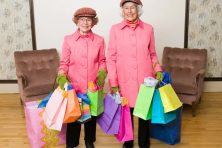 Two senior citizens wear pink coats and a pink hat as they hold multiple shopping bags in their hands.