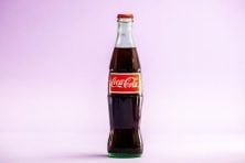 A glass bottle of Coca-Cola is photographed against a magenta background.