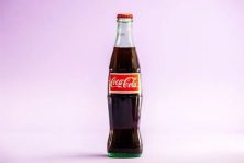 A glass bottle of Coca-Cola is photographed against a magenta background.