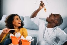 Two people jokingly eat their takeout Chinese food together on the floor in their living room.