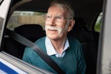 A senior citizen male smiles while driving the backseat of a car with the windows rolled down.