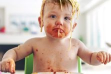 A baby eats spaghetti with pasta all over their head.