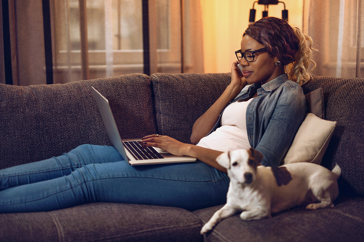 A woman talks on the phone while typing on her laptop and reclining on her couch.