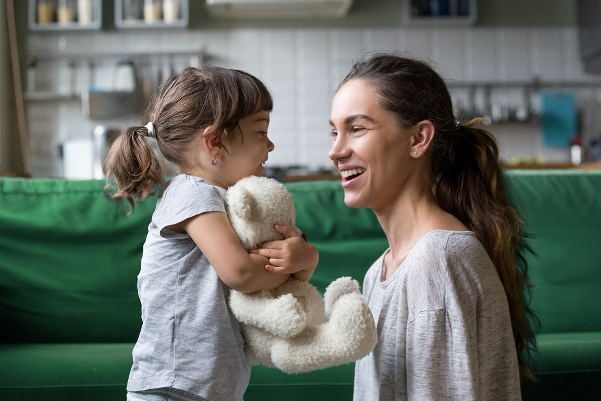 A woman smiles as the child she is watching hugs her bear.