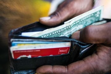 A person holds their wallet open containing dollar bills plus credit cards.