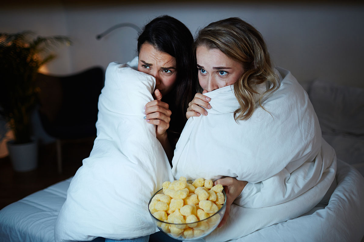 Scared young women hiding in a blanket while watching scary movies at night.