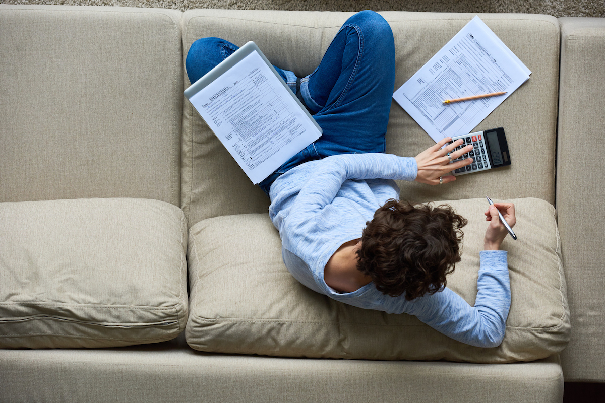 above view of curly-haired woman examining data of tax papers using calculator while sitting on sofa at home