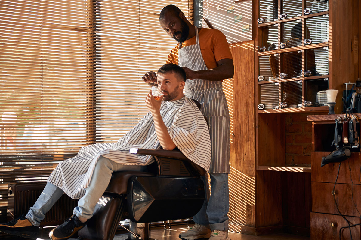 A man gets his hair cut while sipping on a drink. 