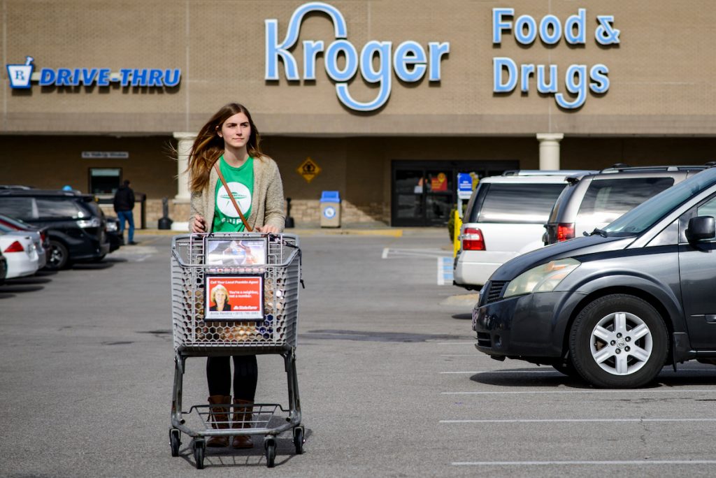 A young woman walks her filled shopping cart. The Kroger storefront can be seen behind her.