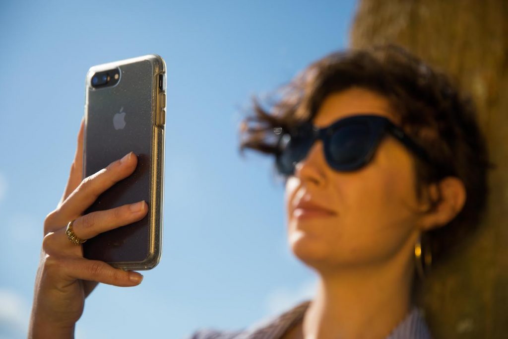 A woman wearing sunglasses leans against a tree as she holds her phone high and close to her face.