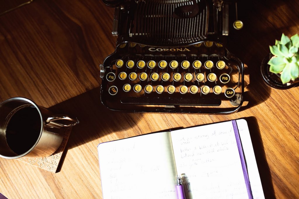 A typewriter sits on top of a desk. Next to the typewriter are a notebook, a succulent, and a cup of coffee.