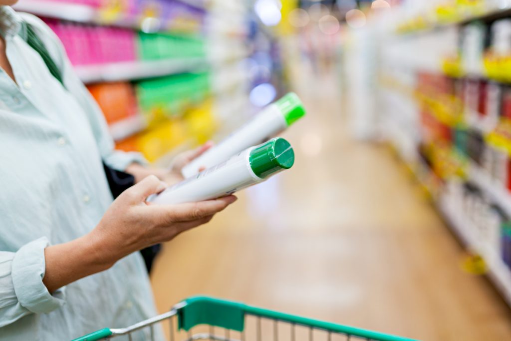 woman in a grocery store aisle holding two shampoo bottles and pushing a shopping cart.