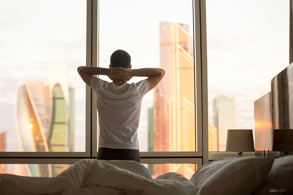 Rear view of young man looking at dawn city scenery in window after waking up.