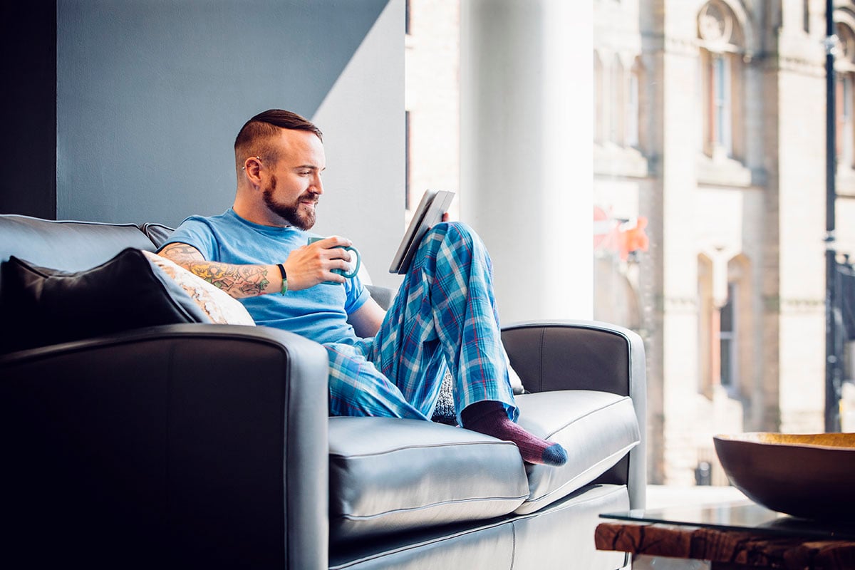 A man in his late 20s is sitting at home on the sofa in his pajamas searching the web on his digital tablet.