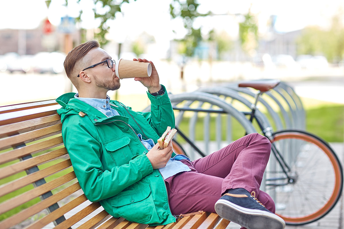 man drinking coffee cup and eating sandwich on city street